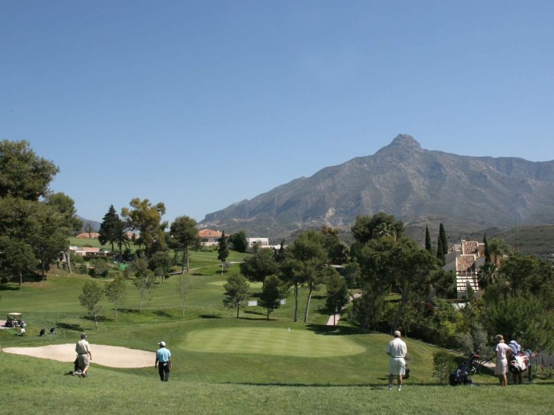 Golfers playing on a lush golf course in Marbella with panoramic views of La Concha mountain, highlighting golf properties in Marbella on the Costa del Sol under a clear blue sky.